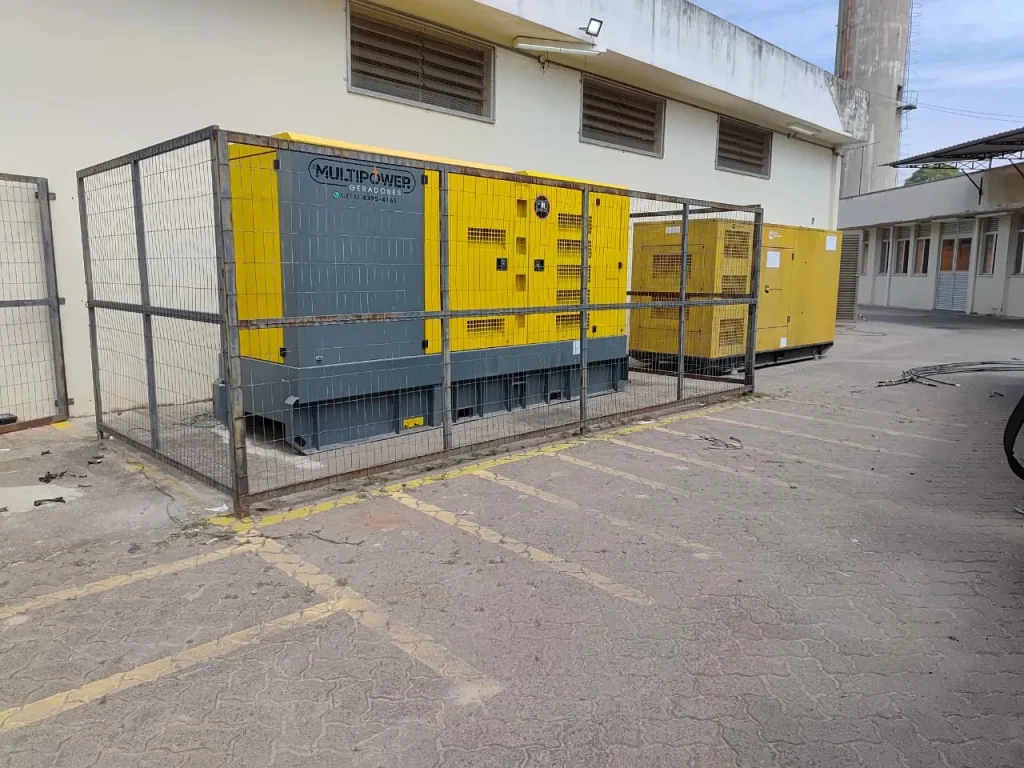 Grupos geradores amarelos protegidos por cerca junto a prédio.